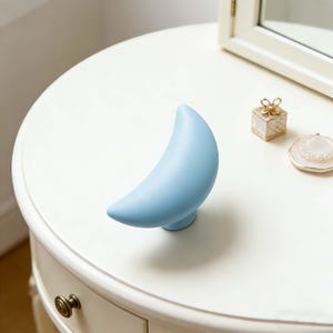 May include: A light blue crescent moon-shaped decorative object on a white table. The moon is smooth and stands upright on a small, round base. Other small decorative items are also on the table.