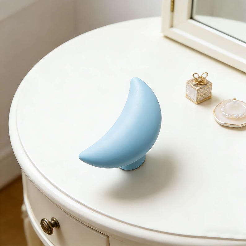 May include: A light blue crescent moon-shaped decorative object on a white table. The moon is smooth and stands upright on a small, round base. Other small decorative items are also on the table.