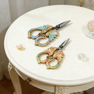 Pode incluir: Duas tesouras decorativas com cabos ornamentados e coloridos. Os cabos apresentam um design de filigrana dourada com detalhes em esmalte e strass em cores pastel. As lâminas são prateadas. As tesouras estão numa mesa branca.