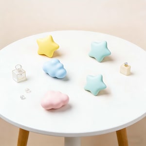 May include: A selection of pastel-coloured decorative items, including yellow stars, blue and pink clouds, and light blue stars, arranged on a white table. Two small perfume bottles and a few clear crystals are also present.