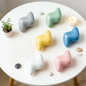Könnte beinhalten: Sieben dekorative Keramikobjekte in verschiedenen Farben, darunter Grau, Grün, Gelb, Blau, Weiß und Rosa. Die Objekte haben eine einzigartige, geschwungene Form und sind auf einem weißen Tisch angeordnet. Eine kleine Topfpflanze und eine brennende Kerze sind ebenfalls vorhanden.