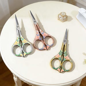 Puede incluir: Tres tijeras de bordado decorativas con diseños florales. Una es plateada con detalles amarillos y verdes, otra es cobriza con detalles rosas y azules, y la tercera es dorada con detalles rosas y verdes. Están dispuestas sobre una superficie blanca.
