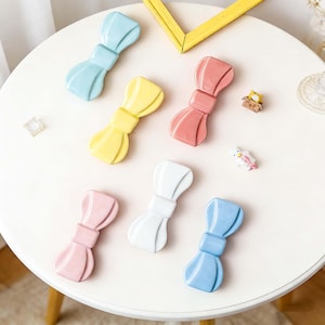 Könnte beinhalten: Eine Sammlung von keramischen Schleifen-Dekorationen in verschiedenen Pastellfarben, darunter Blau, Gelb, Rosa, Weiß und Koralle. Die Schleifen sind auf einem weißen, runden Tisch angeordnet, zusammen mit einem gelben Bilderrahmen und kleinen dekorativen Figuren.