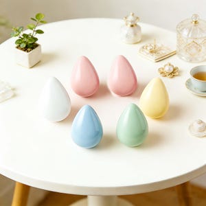May include: Seven teardrop-shaped objects in pastel shades of white, pink, blue, green, and yellow are displayed on a white table. The arrangement suggests a decorative or cosmetic theme, with a focus on colour and form.