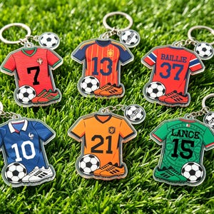 May include: A collection of soccer-themed keychains featuring jersey designs. Each keychain showcases a team jersey with a number, soccer ball, and cleats. The keychains are attached to a silver keyring and a soccer ball charm.