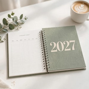 May include: A spiral-bound planner with a sage green cover featuring the year 2027 in large white text. The open page displays a January 2027 calendar grid with columns for days of the week and a checklist section on the left side.