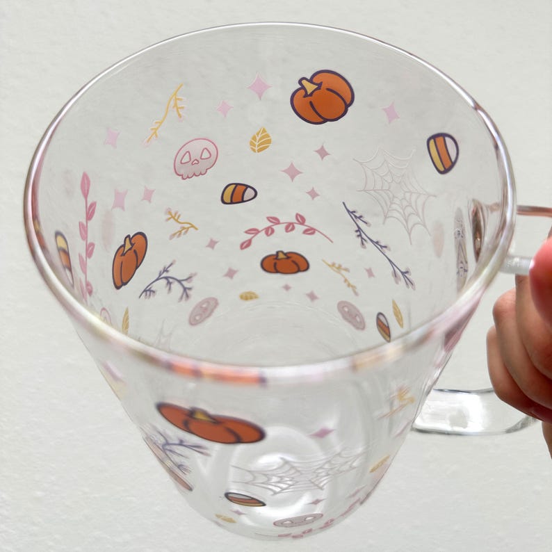 Pode incluir: Uma caneca de vidro transparente com al&ccedil;a, com um design tem&aacute;tico de Halloween. A caneca &eacute; decorada com ab&oacute;boras, milho doce, folhas e teias de aranha em tons de laranja, rosa e amarelo.