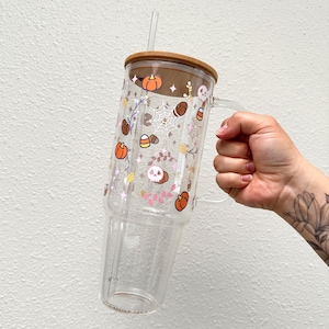 Pode incluir: Um copo de vidro transparente com tampa de madeira e um canudo. O copo &eacute; decorado com um tema de Halloween, com ab&oacute;boras, caveiras, milho doce e folhas em rosa, laranja e marrom.