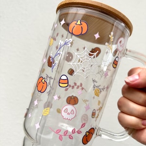 Pode incluir: Uma caneca de vidro transparente com uma tampa de bambu decorada com adesivos tem&aacute;ticos de Halloween. Os adesivos apresentam ab&oacute;boras, milho doce, teias de aranha, caveiras e folhas em rosa, laranja, amarelo e branco.
