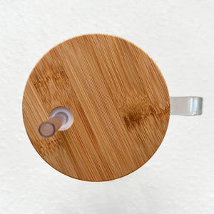 Pode incluir: Uma tampa redonda de madeira com um furo para canudo no centro. A tampa &eacute; feita de bambu marrom claro e tem um padr&atilde;o de gr&atilde;o de madeira natural.