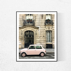 May include: A pink vintage car parked in front of a building with a black door and two balconies with plants. The building has a light beige facade with white trim.