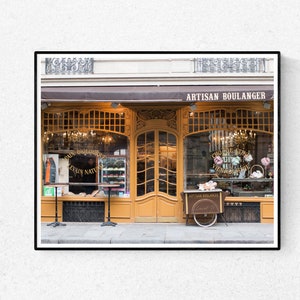 May include: A shop front with a brown awning and gold trim. The sign above the door reads "Artisan Boulanger". The windows are decorated with pastries and flowers. A small cart with pastries is parked in front of the shop.