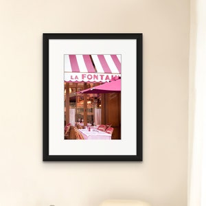 May include: A framed photograph of a Parisian cafe with a pink and white striped awning that reads "LA FONTAINE". The cafe has a pink umbrella and tables set for outdoor dining.