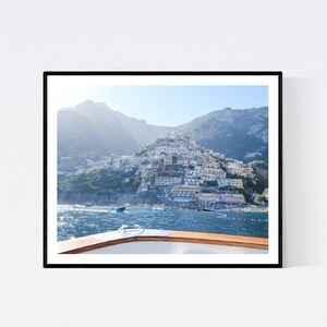 May include: A framed photograph captures a coastal Italian town nestled on a hillside, viewed from a boat. The image features a clear blue sky, the sea, and the town's buildings. The boat's wooden railing is visible in the foreground.