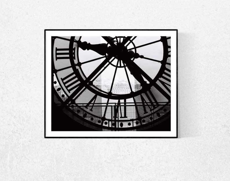 May include: Black and white photograph of a clock face with Roman numerals. The clock is framed in a black frame and the background is a blurry image of a city.