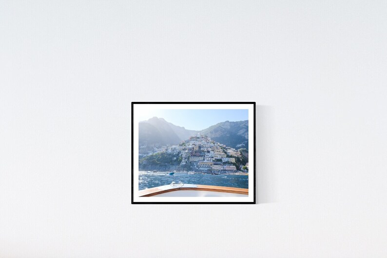 May include: A framed photograph captures a coastal Italian town nestled on a hillside, viewed from a boat. The image features a clear blue sky, the sea, and the town's buildings. The boat's wooden railing is visible in the foreground.