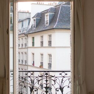 May include: A view from a window with white curtains, looking out at a white building with a slate roof and a wrought iron balcony. The building has several windows with black window frames.