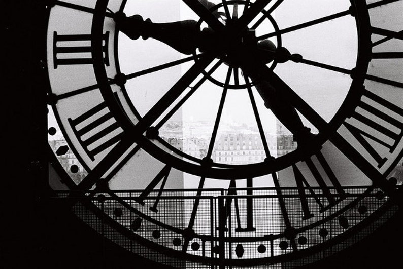 May include: Black and white photo of a large clock face with Roman numerals. The clock face is framed by a metal grid. The clock hands are in silhouette against a bright background.