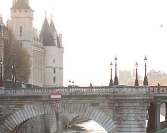 May include: A stone bridge with arches over a river in Paris, France. The bridge has streetlights and a red and white flag on the side. In the background, a tall building with a pointed roof and windows.