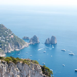 May include: A panoramic view of a rocky island with a town built on the cliffs. The island is surrounded by clear blue water with several boats and yachts. The sky is a light blue with a hazy white background.