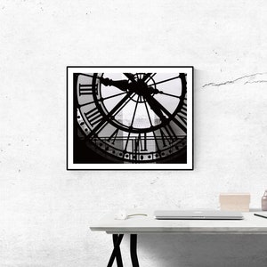 May include: Black and white photograph of a clock face with Roman numerals. The clock is framed in a black frame and is hanging on a white wall.