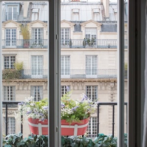 May include: A view from a window of a Parisian apartment building with a balcony and potted plants. The building is made of light-coloured stone with many windows. The balcony is decorated with a red planter box filled with flowers.