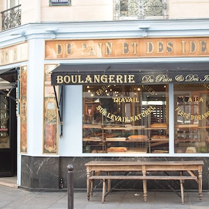 May include: A shop front with a blue awning and the words "Du Pain et Des Idees" in gold lettering. The shop front has a large window display with the words "Boulangerie" and "Du Pain & Des Idees" in gold lettering. The window display features a variety of baked goods, including bread, pastries, and cakes. There is a wooden bench in front of the shop front.