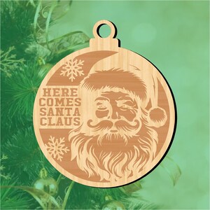 May include: Wooden Christmas bauble with a detailed Santa Claus face, long beard, and hat. The text "HERE COMES SANTA CLAUS" is engraved, along with snowflake designs. The bauble is circular with a hole for hanging, perfect for festive decorations.