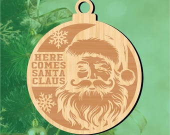 Puede incluir: Adorno navideño de madera con un detallado rostro de Papá Noel, barba larga y gorro. El texto "HERE COMES SANTA CLAUS" está grabado, junto con diseños de copos de nieve. El adorno tiene forma circular con un agujero para colgar.