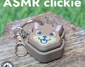 Könnte beinhalten: Ein brauner und beigefarbener ASMR-Clickie-Schlüsselanhänger mit einem Cartoon-Fuchsgesicht-Design. Der Schlüsselanhänger hat einen silbernen Verschluss und einen Ring. Der Text "ASMR clickie" steht in weißen Buchstaben über dem Schlüsselanhänger.