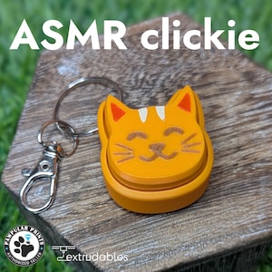 Może przedstawiać: Pomarańczowy brelok w kształcie kota ze srebrnym zapięciem. Kot ma uśmiechniętą twarz z wąsami i białym paskiem na głowie. Tekst "ASMR clickie" znajduje się u góry.