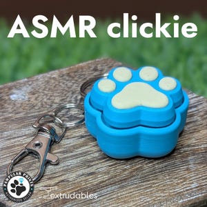 May include: A blue paw-shaped ASMR clickie keychain with a light yellow paw print design. The keychain has a silver clasp and a ring. The text "ASMR clickie" is at the top of the image.