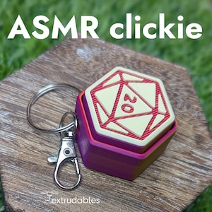 Pode incluir: Um dado hexagonal multicolorido com o número 20 na sua face, preso a um chaveiro. O dado é vermelho, rosa e amarelo. O texto "ASMR clickie" está no topo da imagem. O dado está sobre uma superfície de madeira.
