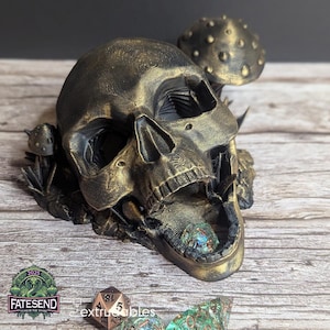 Pode incluir: Um tabuleiro de dados de caveira dourado com um cogumelo no topo. A boca da caveira está aberta e um dado verde e azul está dentro. Há outros dados na superfície de madeira.