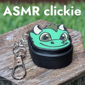 Op de afbeelding: Een zwarte ASMR clickie sleutelhanger met een groen drakenontwerp. De draak heeft witte hoorns en een lachend gezicht. De sleutelhanger heeft een zilveren sluiting en ligt op een houten oppervlak.