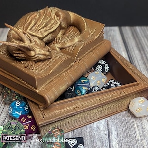 May include: A gold-colored, 3D printed dragon sculpture resting on a closed book. The book is open to reveal a compartment filled with various colored dice. The book is on a wooden surface.