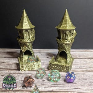 Könnte beinhalten: Zwei goldfarbene 3D-gedruckte Türme mit einer Wendeltreppe, die zu einer kleinen Öffnung an der Spitze führt. Die Türme sind so konzipiert, dass sie Würfel aufnehmen können, und stehen auf einer Holzoberfläche mit mehreren Würfeln, die um sie herum verstreut sind.