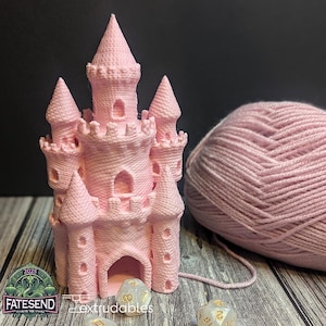 Könnte beinhalten: Ein rosa 3D-gedrucktes Schloss mit einer kleinen Öffnung vorne. Das Schloss steht auf einer Holzoberfläche mit zwei weißen Würfeln davor. Ein Knäuel rosa Garn befindet sich rechts vom Schloss.