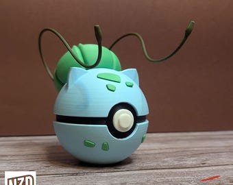 May include: A 3D printed blue and green Pokemon figure of a Bulbasaur. The figure is posed with its arms outstretched and its tail curled up. The figure is sitting on a wooden surface.