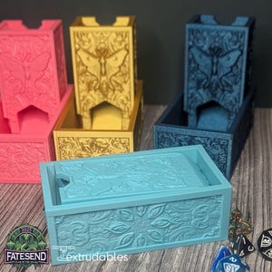 May include: A selection of colourful dice towers and trays. The towers and trays are in shades of pink, gold, and blue. Each tower features a butterfly design. The text "FATESEND YEAR OF THE SNAKE 2025" and "extrudables" are visible.