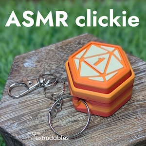 Puede incluir: Un juguete clickie naranja y crema con el texto "ASMR clickie" en la parte superior. El juguete tiene una forma de múltiples lados con el número 1 en la parte superior. Un llavero plateado está unido al juguete.