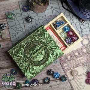 May include: A green book-shaped box with an ornate design and a sword emblem. The box is open, revealing a compartment with dice and a miniature figure.