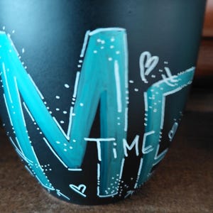 May include: A black ceramic mug with the word "TIME" in white and teal lettering. The letter "M" is stylized with a teal outline and filled in. White dots and a heart shape are also present.