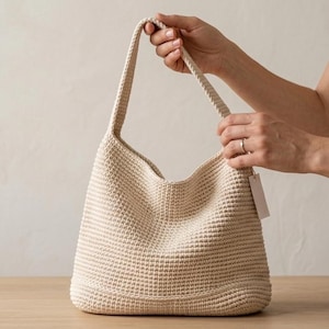 Puede incluir: Un bolso de hombro beige tejido, con una parte superior ancha y abierta y una sola correa. El bolso tiene un patrón texturizado en forma de gofre y lo sostiene una persona. El bolso está sobre una superficie de madera.
