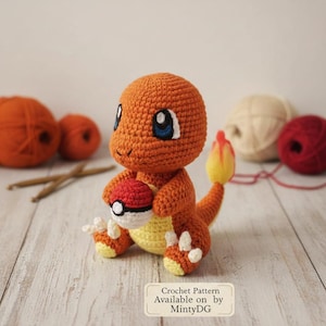 May include: A crocheted orange Charmander figure holding a red and white Poké Ball. The figure has blue eyes, white accents, and a yellow flame tail. The figure is sitting on a white wooden surface. Crochet pattern available.