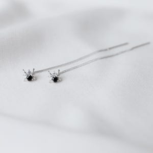 May include: A pair of silver spider-shaped earrings with black gemstone bodies and delicate chain threads. The earrings are displayed on a white, textured surface, creating a minimalist aesthetic.