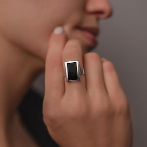 Könnte beinhalten: Ein silberner Ring mit einem rechteckigen schwarzen Stein. Der Ring hat ein schlichtes, modernes Design mit poliertem Finish. Der schwarze Stein ist in eine silberne Fassung gefasst.