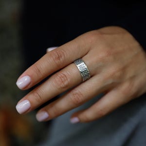 Puede incluir: Un anillo de plata con un patrón geométrico grabado y un emblema central. El anillo es ancho y se lleva en un dedo. El diseño incluye un patrón repetitivo y un emblema detallado.