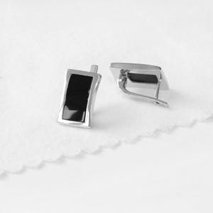 Puede incluir: Un par de pendientes de plata con inserciones rectangulares negras. Los pendientes tienen un diseño simple y moderno.