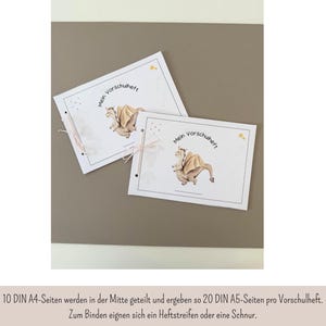 May include: Two white notebooks with the text "Mein Vorschuheft" and a cartoon dragon illustration. The notebooks are bound with a pink ribbon. The background is a solid, neutral color.